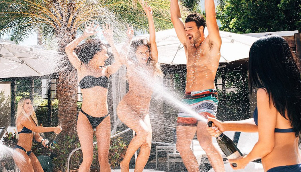 Cheers and laughter fill the air as champagne bottles spray in a playful battle at the Fontainebleau pool in Miami Beach, creating a sparkling and joyous spectacle amidst the glamorous poolside setting.