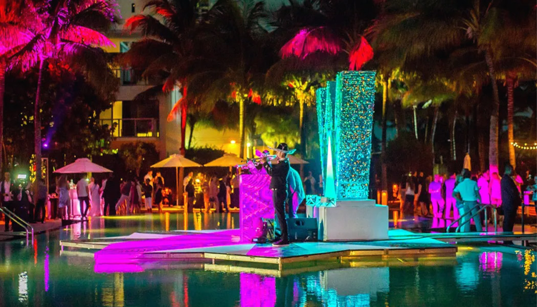 An opulent scene unfolds at the W South Beach Wall Club, with an exclusive poolside party exuding top-class elegance. Guests in chic attire mingle by the pool, enjoying a night of sophistication and luxury, under the starry Miami sky.