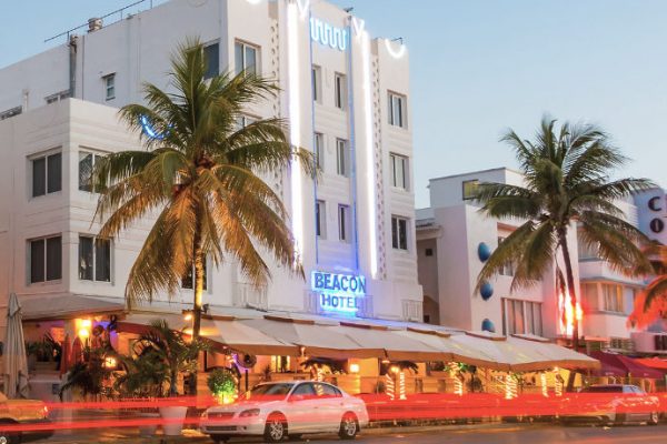 The image captures the brightly lit Beacon South Beach Hotel in Miami's South Beach district at night, with its colorful Art Deco facade and palm trees creating a tropical and luxurious atmosphere.