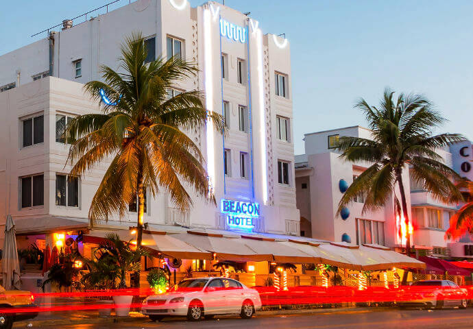 The image captures the brightly lit Beacon South Beach Hotel in Miami's South Beach district at night, with its colorful Art Deco facade and palm trees creating a tropical and luxurious atmosphere.